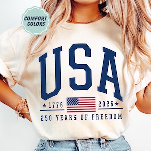 May include: Cream-colored sweatshirt with the word "USA" in navy blue block letters. Below the text is a small American flag and the text "1776" and "2026" with stars. The phrase "250 YEARS OF FREEDOM" is printed below.