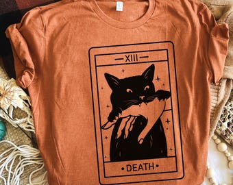 Puede incluir: Una camiseta naranja con un gráfico negro de un gato sosteniendo una mano, enmarcado en un diseño de carta de tarot. La carta dice "-XIII- DEATH-". La camiseta está hecha de un material suave y tiene un cuello redondo clásico.