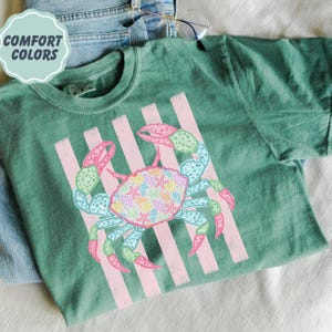 May include: Green t-shirt with a crab graphic. The crab is decorated with seashells and starfish, and has pink and blue accents. The shirt also features pink vertical stripes and the text "COMFORT COLORS" in a light blue circle.