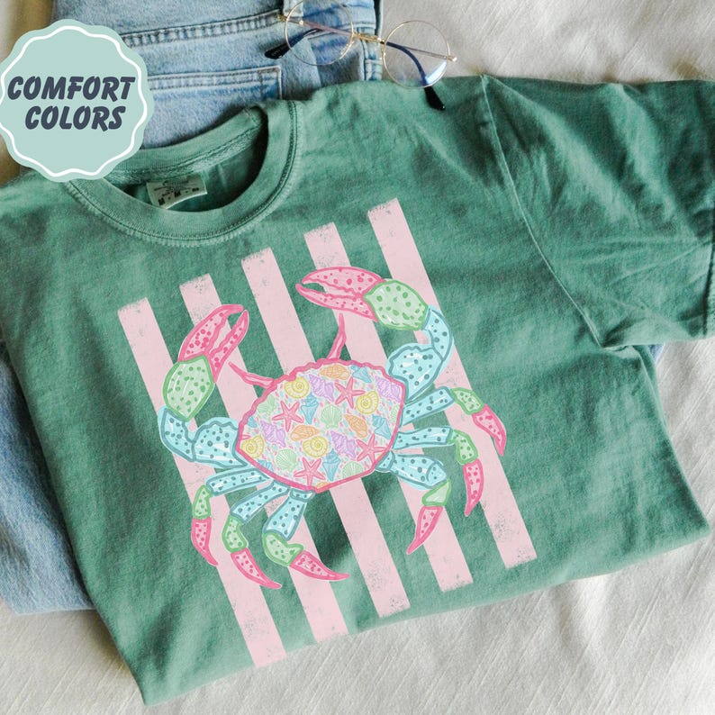 May include: Green t-shirt with a crab graphic. The crab is decorated with seashells and starfish, and has pink and blue accents. The shirt also features pink vertical stripes and the text "COMFORT COLORS" in a light blue circle.