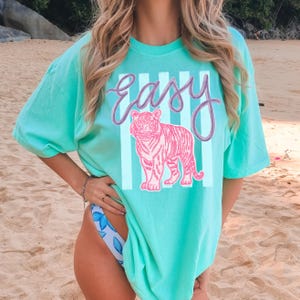 K&ouml;nnte beinhalten: T&uuml;rkisfarbenes T-Shirt mit einem rosa Tiger-Design und dem Wort "Easy" in Schreibschrift. Das Design enth&auml;lt wei&szlig;e vertikale Streifen. Das Shirt hat einen l&auml;ssigen, &uuml;bergro&szlig;en Stil.