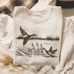 K&ouml;nnte beinhalten: Cremefarbenes Sweatshirt mit einer detaillierten Illustration von zwei Enten im Flug &uuml;ber Wasser mit Schilf. Die Enten haben braune, gr&uuml;ne und wei&szlig;e Markierungen. Das Design hat ein Natur- oder Wildtier-Thema.