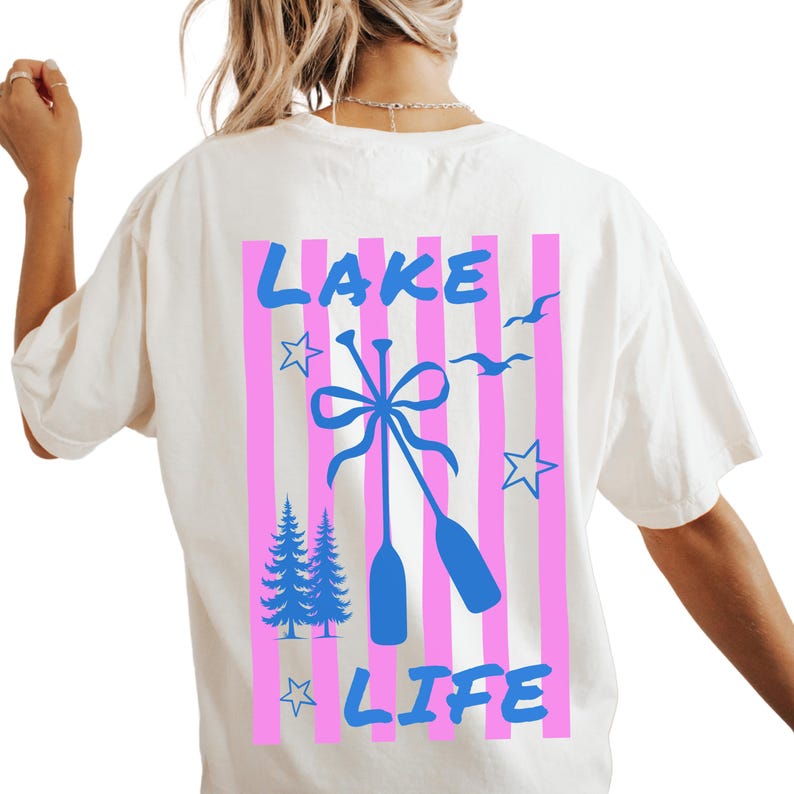 May include: Cream-colored t-shirt with a graphic design on the back. The design features the words "LAKE LIFE" in blue, with pink vertical stripes, blue oars, stars, pine trees, and birds.