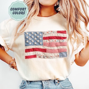 K&ouml;nnte beinhalten: Cremefarbenes Sweatshirt mit einem Patchwork-Design der amerikanischen Flagge. Die Flagge hat blaue Sterne, rote Streifen und Blumen- und Vichy-Muster. Der Text "COMFORT COLORS" befindet sich in einem hellblauen Kreis.