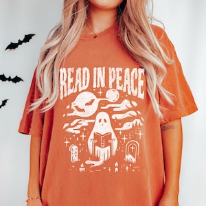 May include: Burnt orange t-shirt with a white graphic that reads "READ IN PEACE." The graphic features a ghost reading a book, bats, clouds, stars, and tombstones. The shirt has short sleeves and a crew neck.
