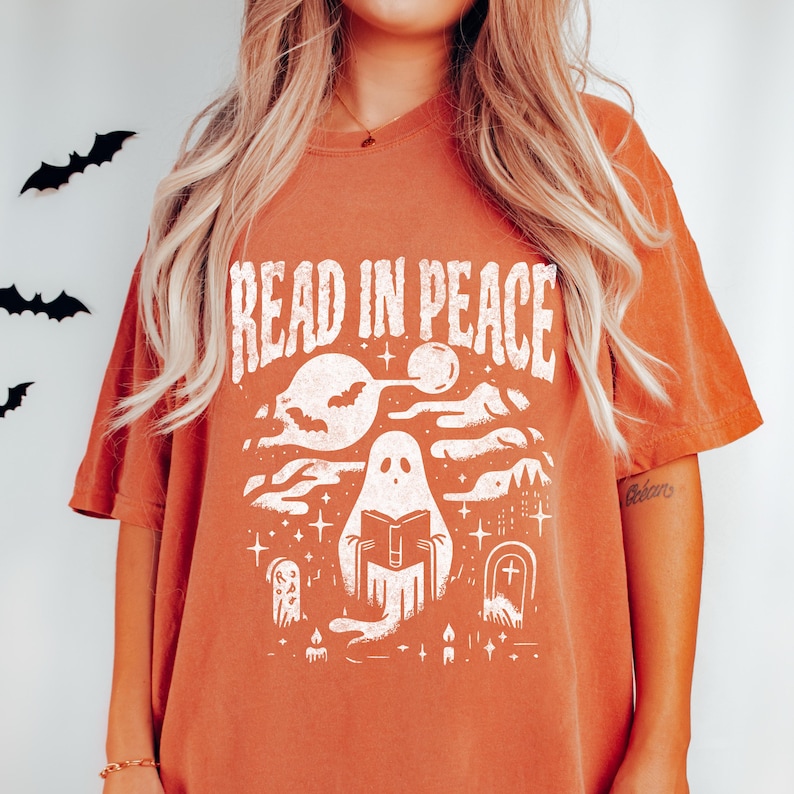 May include: Burnt orange t-shirt with a white graphic that reads "READ IN PEACE." The graphic features a ghost reading a book, bats, clouds, stars, and tombstones. The shirt has short sleeves and a crew neck.