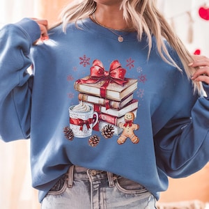 May include: A blue sweatshirt with a Christmas-themed graphic. The graphic depicts a stack of books with a red bow, a mug of whipped cream, a gingerbread man, pine cones, and snowflakes. The sweatshirt is a solid blue colour.