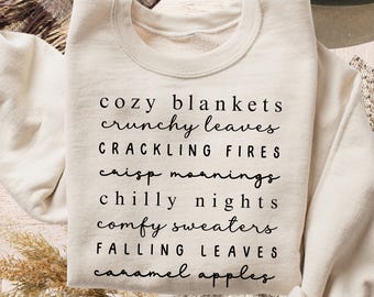 May include: Cream-colored sweatshirt with black text listing autumn-themed phrases: "cozy blankets," "crunchy leaves," "crackling fires," "crisp mornings," "chilly nights," "comfy sweaters," "falling leaves," and "caramel apples."