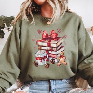 May include: An olive green sweatshirt with a festive graphic. The graphic depicts a stack of books tied with a red bow, a mug of whipped cream, a gingerbread man, pine cones, and snowflakes. The sweatshirt is a casual, comfortable style.