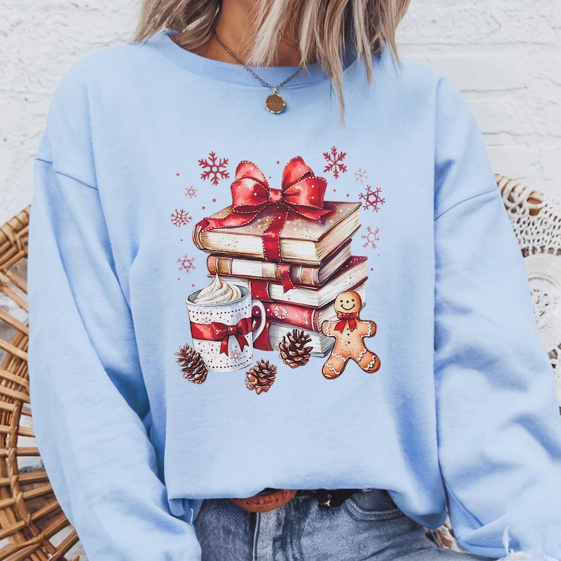 May include: Pale blue sweatshirt with a festive illustration. A stack of books tied with a red bow, a mug of whipped cream, a gingerbread man, snowflakes, and pine cones. The design evokes a cosy, holiday theme.