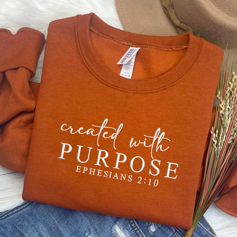 May include: An orange crew neck sweatshirt with the words "created with PURPOSE" and "EPHESIANS 2:10" printed in white. The sweatshirt is folded on top of blue denim jeans. A Jerzees brand tag is visible.