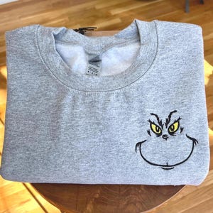 Puede incluir: Una sudadera gris con la cara del Grinch bordada en la parte delantera. La cara del Grinch est&aacute; delineada en negro con ojos amarillos. La sudadera est&aacute; doblada y parece estar hecha de un material suave.