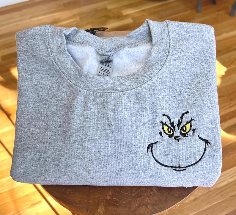 Puede incluir: Una sudadera gris con la cara del Grinch bordada en la parte delantera. La cara del Grinch est&aacute; delineada en negro con ojos amarillos. La sudadera est&aacute; doblada y parece estar hecha de un material suave.