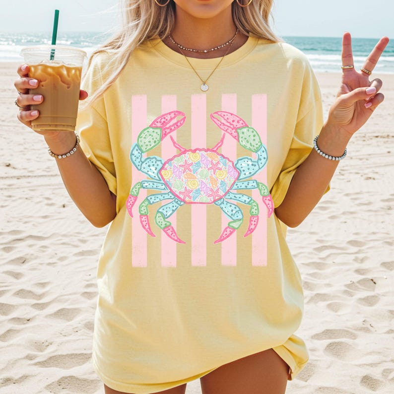 May include: Yellow t-shirt featuring a colorful crab design with pink and white stripes. The crab has blue, green, and pink accents, and is filled with seashell patterns. The shirt is a casual, beach-themed apparel item.