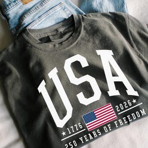 May include: Dark gray t-shirt with the word "USA" in large white letters. Below the text is a small American flag and the text "1776 2026 250 YEARS OF FREEDOM". Light blue denim jeans are folded behind the shirt.