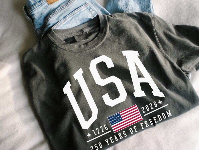 May include: Dark gray t-shirt with the word "USA" in large white letters. Below the text is a small American flag and the text "1776 2026 250 YEARS OF FREEDOM". Light blue denim jeans are folded behind the shirt.