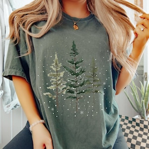 May include: Dark green t-shirt featuring a watercolor design of three evergreen trees with falling snow. The trees are in shades of green and white, creating a winter or holiday theme. The shirt has a relaxed fit and short sleeves.