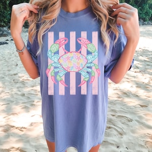 May include: A blue t-shirt featuring a colorful crab design with pink and white stripes. The crab has a variety of colors, including pink, green, blue, and yellow. The shirt is a comfortable, oversized fit.