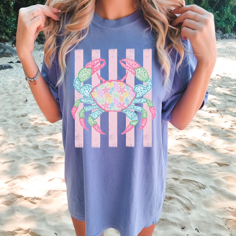 May include: A blue t-shirt featuring a colorful crab design with pink and white stripes. The crab has a variety of colors, including pink, green, blue, and yellow. The shirt is a comfortable, oversized fit.
