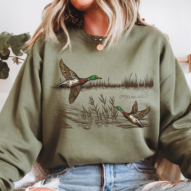 K&ouml;nnte beinhalten: Ein olivgr&uuml;nes Sweatshirt mit einer Grafik von zwei Enten im Flug &uuml;ber Wasser, mit Schilf im Hintergrund. Die Enten sind braun, wei&szlig; und gr&uuml;n. Das Sweatshirt ist ein l&auml;ssiger Stil.