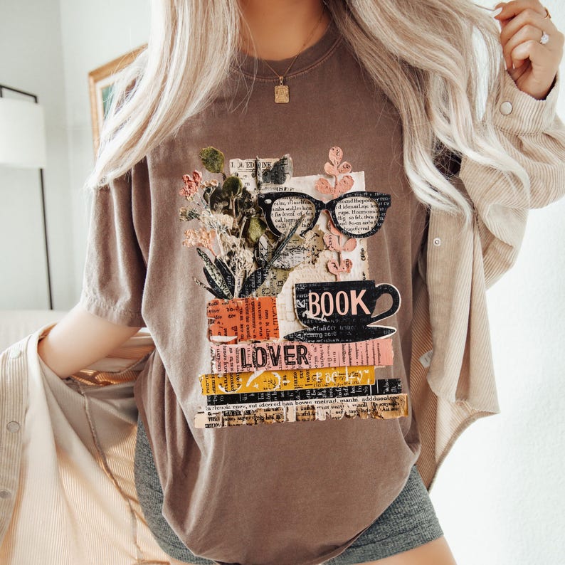 May include: Brown t-shirt with a graphic design featuring a stack of books, eyeglasses, and flowers. The word "BOOK" is printed on a teacup, and the word "LOVER" is printed on the books. The shirt has a relaxed fit.