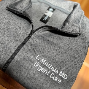 May include: A gray, long-sleeved athletic jacket with a black zipper and a mock neck collar. The jacket has the embroidered text "L. Mallinis MD Urgent Care" in white. A tag with a logo is visible.