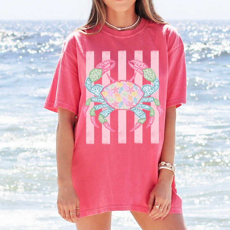 May include: A coral-colored t-shirt featuring a colorful crab graphic. The crab is centered on the shirt, with a striped background. The crab design incorporates pastel colors, including blue, green, pink, and yellow, creating a beachy theme.