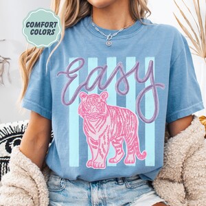 K&ouml;nnte beinhalten: Blaues T-Shirt mit dem Wort "Easy" in Schreibschrift und einer rosa Tigergrafik. Hinter dem Text und dem Tiger befinden sich hellblaue vertikale Streifen. Die Worte "Comfort Colors" stehen in einem hellgr&uuml;nen Kreis oben links.
