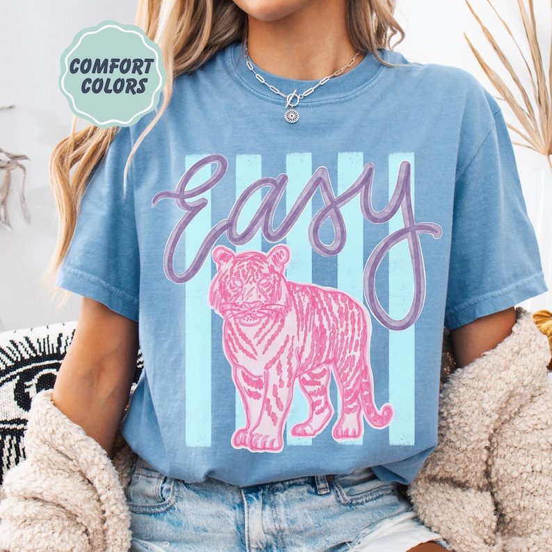 K&ouml;nnte beinhalten: Blaues T-Shirt mit dem Wort "Easy" in Schreibschrift und einer rosa Tigergrafik. Hinter dem Text und dem Tiger befinden sich hellblaue vertikale Streifen. Die Worte "Comfort Colors" stehen in einem hellgr&uuml;nen Kreis oben links.