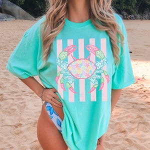 May include: A turquoise t-shirt featuring a colorful crab design with pink and green accents. The crab is centered on a background of vertical pink and white stripes. The shirt is a comfortable, oversized fit.