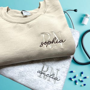 Puede incluir: Dos sudaderas beige y gris con las letras RN bordadas. La sudadera beige tiene el nombre "sophia" bordado debajo de las letras. La sudadera gris tiene el nombre "amelia" bordado debajo de las letras.