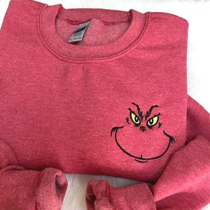 Puede incluir: Una sudadera roja con cuello redondo y mangas largas. La sudadera presenta una cara del Grinch bordada con ojos amarillos y un contorno negro. La tela tiene una textura jaspeada.