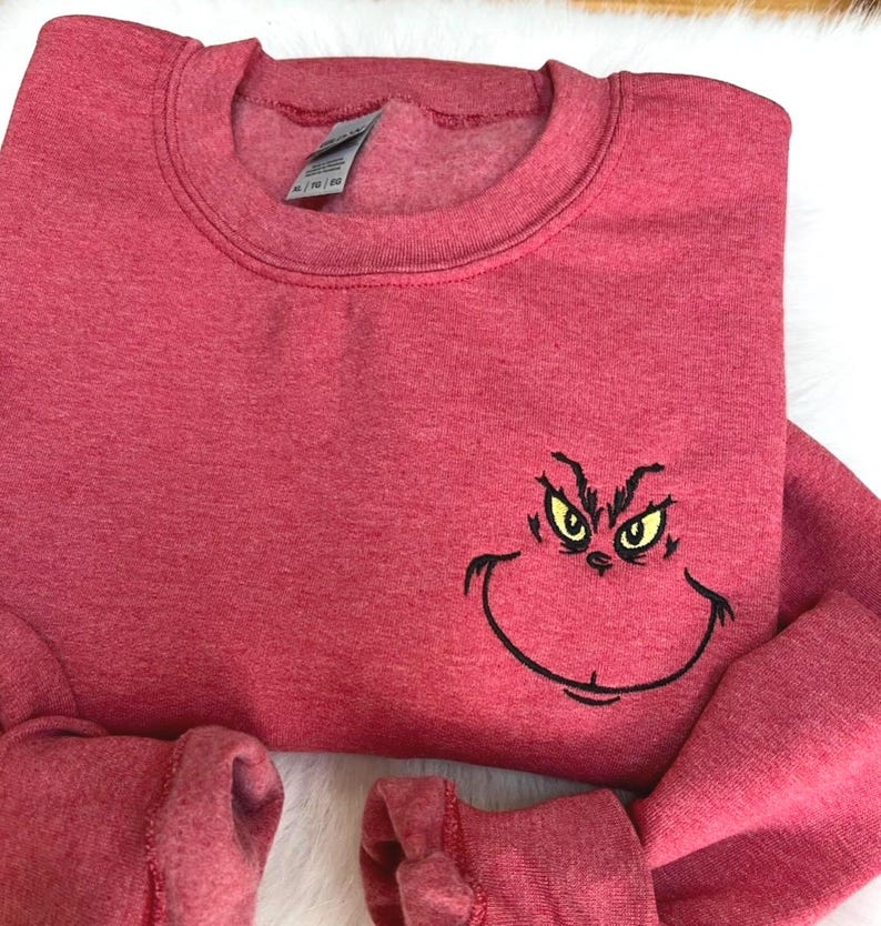 Puede incluir: Una sudadera roja con cuello redondo y mangas largas. La sudadera presenta una cara del Grinch bordada con ojos amarillos y un contorno negro. La tela tiene una textura jaspeada.
