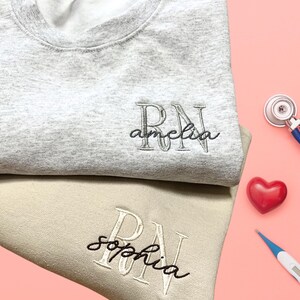 Puede incluir: Dos sudaderas grises y beige con las letras "RN" bordadas. Las sudaderas tambi&eacute;n tienen los nombres "Amelia" y "Sophia" bordados.