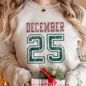 Pu&ograve; includere: Felpa color crema con la scritta "DECEMBER 25" in un carattere invecchiato in stile collegiale. Il testo &egrave; in bordeaux e verde. La felpa fa parte di una collezione a tema festivo.