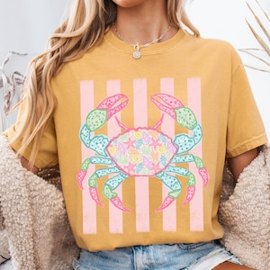 May include: Mustard yellow t-shirt featuring a colorful crab design. The crab is decorated with seashells and starfish, set against pink vertical stripes. The crab's claws and legs are in shades of pink, green, and blue.