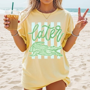 Puede incluir: Una camiseta amarilla con la palabra "later" en verde sobre una ilustraci&oacute;n de un caim&aacute;n verde. La camiseta tiene un fondo a rayas y se usa en la playa.