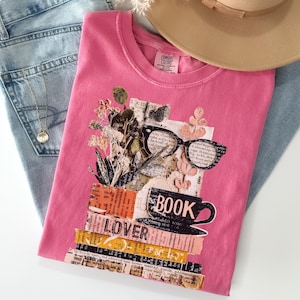 May include: A pink t-shirt with a collage design featuring glasses, a coffee cup, and the word "BOOK". The design includes the word "LOVER" and floral elements. The shirt is folded on top of blue jeans.
