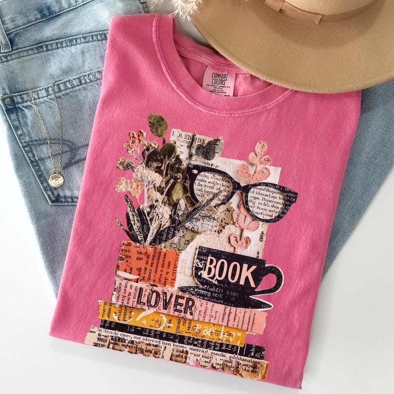 May include: A pink t-shirt with a collage design featuring glasses, a coffee cup, and the word "BOOK". The design includes the word "LOVER" and floral elements. The shirt is folded on top of blue jeans.