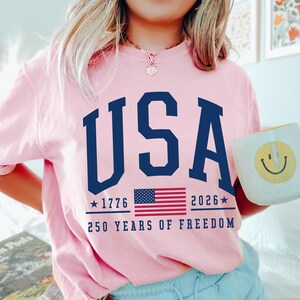 May include: Pink sweatshirt with the word "USA" in navy blue block letters. Below the word "USA" is the American flag, the years "1776" and "2026", and the text "250 YEARS OF FREEDOM". A white mug with a yellow smiley face is held in the hand.