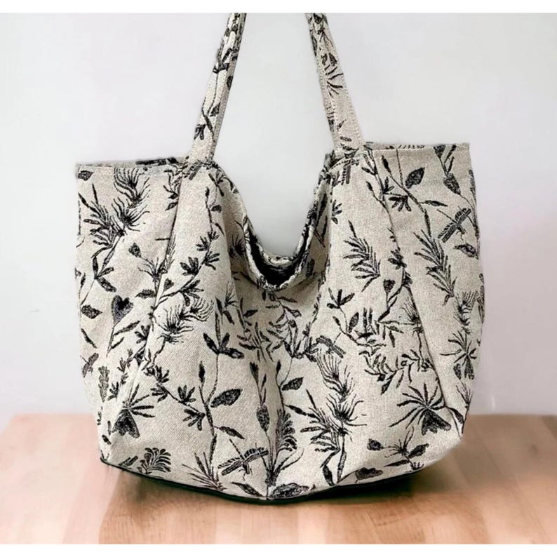 May include: A large, beige tote bag with black floral print. The bag has two long handles and a wide opening.