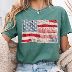 K&ouml;nnte beinhalten: Ein teal-farbenes T-Shirt mit einem Patchwork-Design der amerikanischen Flagge. Die Flagge verwendet Stoffe mit roten, wei&szlig;en und blauen Mustern und gestickten Sternen. Das T-Shirt hat kurze &Auml;rmel und eine lockere Passform, geeignet f&uuml;r Freizeitkleidung.