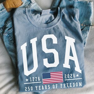 May include: A blue t-shirt with the word "USA" in large white letters. Below the text is a small American flag and the text "1776 2026" and "250 YEARS OF FREEDOM". The shirt is folded on top of a pair of blue jeans.