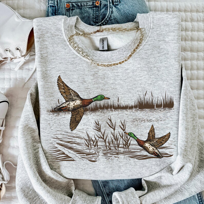 K&ouml;nnte beinhalten: Ein hellgraues Sweatshirt mit einer detaillierten Illustration von zwei Enten im Flug &uuml;ber Wasser mit Schilf. Die Enten haben braune, gr&uuml;ne und wei&szlig;e Markierungen. Eine goldene Halskette ist auf dem Sweatshirt drapiert.