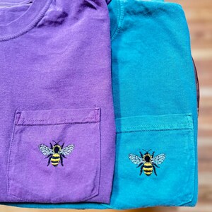 May include: Two short-sleeved t-shirts, one purple and one teal, each featuring a chest pocket. An embroidered graphic of a bumblebee with yellow and black stripes and light blue wings is centered on the front of each pocket on both shirts.