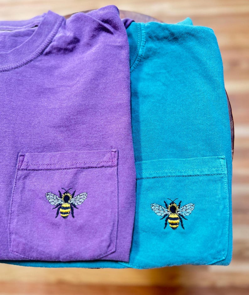 May include: Two short-sleeved t-shirts, one purple and one teal, each featuring a chest pocket. An embroidered graphic of a bumblebee with yellow and black stripes and light blue wings is centered on the front of each pocket on both shirts.