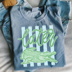 Puede incluir: Una camiseta azul con la palabra "later" en verde, sobre un fondo de rayas verticales azul claro. Debajo, una ilustraci&oacute;n de un caim&aacute;n verde. La camiseta est&aacute; doblada sobre unos vaqueros azules.