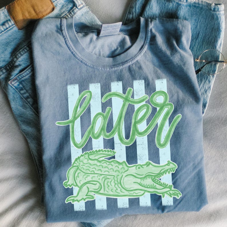 Puede incluir: Una camiseta azul con la palabra "later" en verde, sobre un fondo de rayas verticales azul claro. Debajo, una ilustraci&oacute;n de un caim&aacute;n verde. La camiseta est&aacute; doblada sobre unos vaqueros azules.