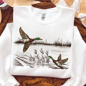 K&ouml;nnte beinhalten: Wei&szlig;es Sweatshirt mit einer detaillierten Illustration von zwei Enten im Flug &uuml;ber Wasser und Schilf. Die Enten sind braun, wei&szlig; und gr&uuml;n. Das Sweatshirt ist von der Marke Gildan.
