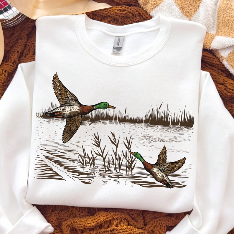 K&ouml;nnte beinhalten: Wei&szlig;es Sweatshirt mit einer detaillierten Illustration von zwei Enten im Flug &uuml;ber Wasser und Schilf. Die Enten sind braun, wei&szlig; und gr&uuml;n. Das Sweatshirt ist von der Marke Gildan.